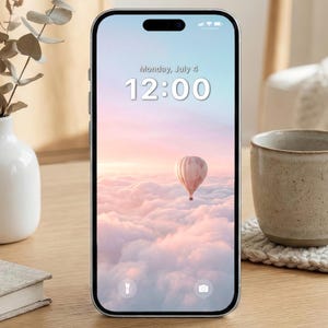 May include: A smartphone displays a pastel-colored wallpaper featuring a hot air balloon floating above fluffy clouds. The screen shows the date as Monday, July 4, and the time as 12:00. The phone is on a wooden surface.