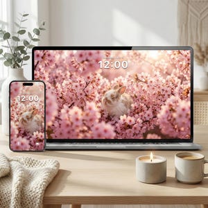 May include: A laptop and smartphone display a wallpaper of a bunny nestled in pink cherry blossoms. The time, 12:00, is displayed on both screens. A lit candle and a mug of coffee sit on a wooden table, alongside a vase with greenery and a folded blanket.
