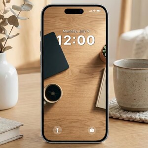 May include: A smartphone displays a wooden desk scene with a black notebook, a cup of coffee, a small plant, and a book. The screen shows the date as Monday, July 4, and the time as 12:00. A white vase and a ceramic mug are also visible.