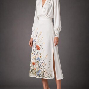 May include: A white midi dress featuring a mock neck, long sleeves with buttoned cuffs, and a wrap-style bodice. The skirt includes a side slit and a colorful botanical print of wildflowers, including red poppies and blue cornflowers, along the hem.