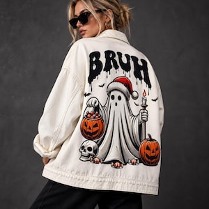 Puede incluir: Una chaqueta de mezclilla color hueso con un gráfico grande en la espalda de un fantasma con gorro de Santa, sosteniendo una vela encendida y un cubo de calabaza lleno de dulces. El texto "BRUH" aparece en negro sobre el fantasma, con una calavera.
