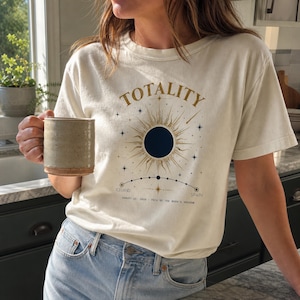 Peut inclure: Un t-shirt à manches courtes de couleur crème arborant un graphique d'éclipse solaire avec le mot TOTALITY en lettres dorées. Le motif inclut des étoiles et une trajectoire de l'ombre de la lune marquée ICELAND et SPAIN.