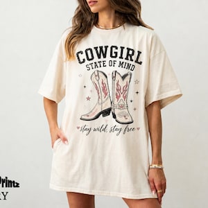 Peut inclure: T-shirt ivoire avec l'inscription "COWGIRL STATE OF MIND" au-dessus d'un motif de bottes de cowboy. Les bottes sont roses et rouges avec des étoiles. Le texte "stay wild, stay free" est en dessous.