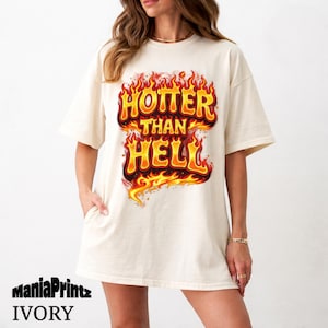 Peut inclure: T-shirt ivoire avec l'inscription "HOTTER THAN HELL" dans un design flamboyant orange et jaune. Le t-shirt a des manches courtes et une coupe ample. La marque "ManiaPrintz" et la couleur "IVORY" sont imprimées en bas.
