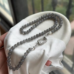 Puede incluir: Un collar de cuentas grises con un cierre plateado. El collar está hecho de cuentas redondas grises y tiene un cierre plateado con pequeñas piedras transparentes. Se muestra sobre una tela blanca.