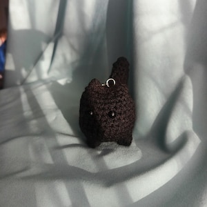 Może przedstawiać: Mała, ciemnobrązowa figurka kota zrobiona na szydełku, z czarnymi koralikowymi oczami i małym ogonem. Z tyłu przymocowana jest srebrna metalowa pętla, prawdopodobnie do breloczka lub ozdoby. Tło to miękka, jasnoniebieska tkanina.