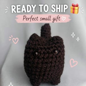 Può includere: Un peluche a forma di gatto all'uncinetto marrone scuro con occhi a bottone. Il testo "READY TO SHIP" e "Perfect small gift" sono visualizzati sopra il peluche. Sono inclusi anche cuori rosa e una grafica a forma di confezione regalo.