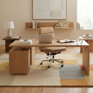 May include: A contemporary home office with a wooden desk, laptop, and leather chair. A colourful geometric rug and side table with a lamp are also present. The room is decorated with a potted plant and artwork.