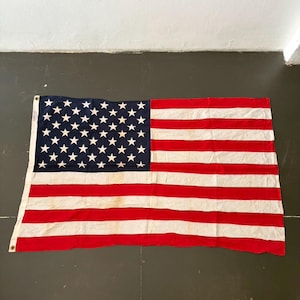 May include: A vintage American flag, with a blue canton filled with white stars and red and white stripes. The rectangular flag is made of fabric. The flag is displayed on a dark surface.