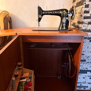 Op de afbeelding: Een vintage Singer naaimachine, zwart met gouden accenten, staat op een houten kast. De kast heeft een lade met spoelen en naai-accessoires. Het wiel van de machine en andere onderdelen zijn zichtbaar.