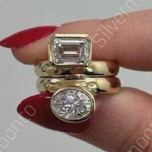 May include: Three gold rings with clear gemstones. The top ring has an emerald-cut stone, the middle ring is plain, and the bottom ring has an oval-cut stone. The rings are stacked together.