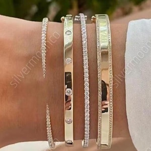 Puede incluir: Una colección de pulseras de oro y plata. Las pulseras incluyen una pulsera de tenis con diamantes, una pulsera de oro con piedras transparentes y una pulsera de oro con una superficie lisa. Las pulseras se llevan en la muñeca.