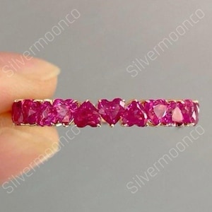 Op de afbeelding: Een gouden ring met een rij hartvormige, helder roze edelstenen. De edelstenen zijn dicht op elkaar geplaatst, waardoor een doorlopende band van kleur ontstaat. De ring wordt tegen een neutrale achtergrond gehouden.