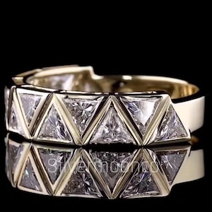 May include: A gold band ring featuring a row of triangular-cut clear stones set in a repeating geometric pattern. The metal has a polished finish, and the design includes a series of triangular bezels that hold each stone securely in place.
