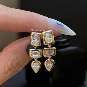 May include: Gold-toned drop earrings with four clear, sparkling gemstones. The earrings feature a rectangular top, a rectangular middle, and a pear-shaped bottom. The earrings are held up by a hand with a manicure. The text "Silvermoonco" is visible.