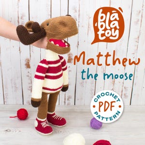 Puede incluir: Un juguete de ganchillo con forma de alce llamado "Matthew", con cuernos, cabeza y patas marrones, que lleva un jersey a rayas rojas y blancas y zapatillas rojas. La imagen incluye el texto "bla toy" y "Matthew the moose". También se ve un "Crochet PDF Pattern".
