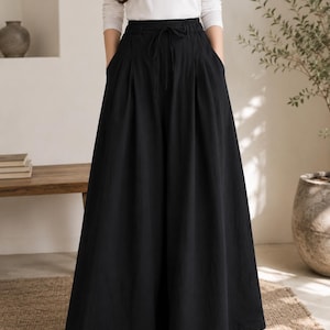 May include: High-waisted black wide-leg trousers featuring a gathered elastic waistband with a matching drawstring tie, deep side pockets, and a floor-length, voluminous silhouette that flares out from the hips to the hemline.