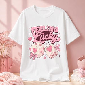 May include: A white short-sleeved t-shirt featuring a graphic print with the text "Feeling Lucky" in pink bubble letters. The design includes two light pink dice with heart-shaped pips, a pink bow, small stars, and a smiling face icon.