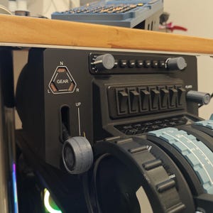 May include: Black flight simulator control panel with various switches, dials, and levers. The panel includes a gear indicator, labelled "GEAR," and a lever marked "UP." The design features a mix of grey and black components, suggesting a realistic cockpit setup.