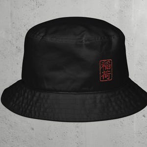 May include: A solid black bucket hat featuring a small, rectangular red embroidered patch on the side. The embroidery depicts two Japanese kanji characters enclosed within a thin red border, creating a distinct contrast against the dark fabric.