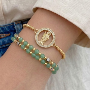 May include: Two bracelets on a wrist. One is gold beaded with a circular pendant featuring a religious figure and clear stones. The other bracelet has green beads and gold and silver accents. The bracelets are worn on a wrist.