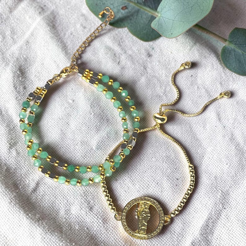May include: Two gold-toned bracelets. One features small, round, light green beads and gold accents. The other has a gold chain with a circular pendant with a figure inside. The bracelets are on a white surface.