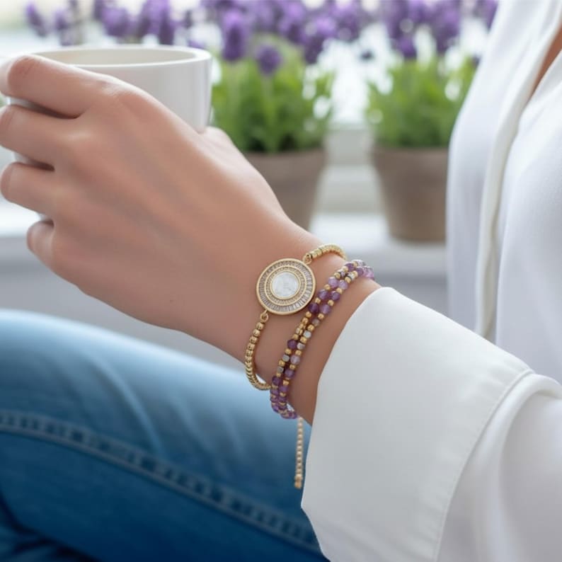 May include: A gold bracelet with a round, white center stone and a smaller gold beaded bracelet with purple beads. The bracelets are stacked on a wrist, with a white shirt and blue jeans in the background.