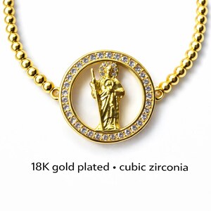 May include: Gold-plated necklace featuring a central pendant with a religious figure encircled by cubic zirconia stones. The necklace has a beaded chain and the text "18K gold plated &bull; cubic zirconia".