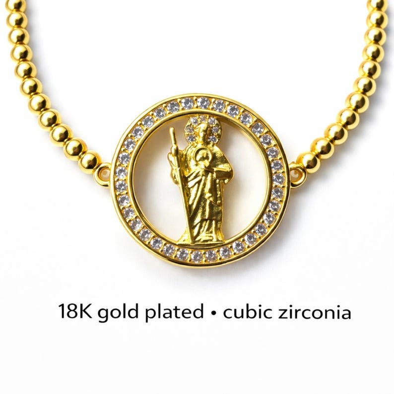 May include: Gold-plated necklace featuring a central pendant with a religious figure encircled by cubic zirconia stones. The necklace has a beaded chain and the text "18K gold plated &bull; cubic zirconia".