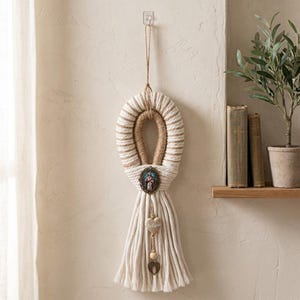 May include: A wall hanging featuring a looped cream and tan rope design with a central oval religious medallion depicting a figure. Below the medallion, a dangling charm includes a heart-shaped bead with the word Believe and a small metal wing charm.