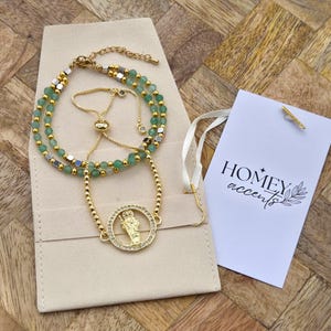 May include: Two gold-colored bracelets with green beads and a circular pendant with a religious figure. One bracelet is adjustable. The bracelets are on a beige pouch, with a tag that reads "HOMEY accents".