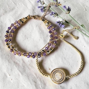 May include: Two bracelets. One features amethyst beads and gold-colored accents. The other is a gold-colored adjustable bracelet with a round pendant. The pendant has a central design surrounded by small crystals. Lavender sprigs are in the background.