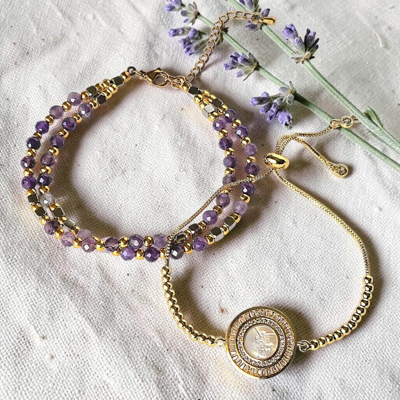 May include: Two bracelets. One features amethyst beads and gold-colored accents. The other is a gold-colored adjustable bracelet with a round pendant. The pendant has a central design surrounded by small crystals. Lavender sprigs are in the background.