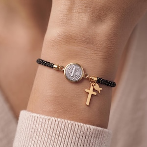 Puede incluir: Una pulsera de cordón trenzado negro con una medalla de San Benito plateada en el centro, engastada en un marco dorado. Dos dijes dorados, una cruz sencilla y un pequeño símbolo de pez, cuelgan del lateral del engaste central.