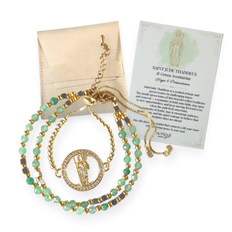 May include: A gold-colored bracelet with green beads and a circular pendant featuring a saint figure. The bracelet is accompanied by a card with text about Saint Jude Thaddeus and green aventurine. A small beige pouch is also included.