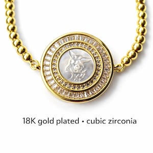 May include: Gold-plated bracelet with a round pendant featuring a carved design and cubic zirconia accents. The pendant has a central design and is surrounded by a ring of clear stones. The bracelet is made of small, gold-colored beads. Text on the image reads: "18K gold plated &bull; cubic zirconia."