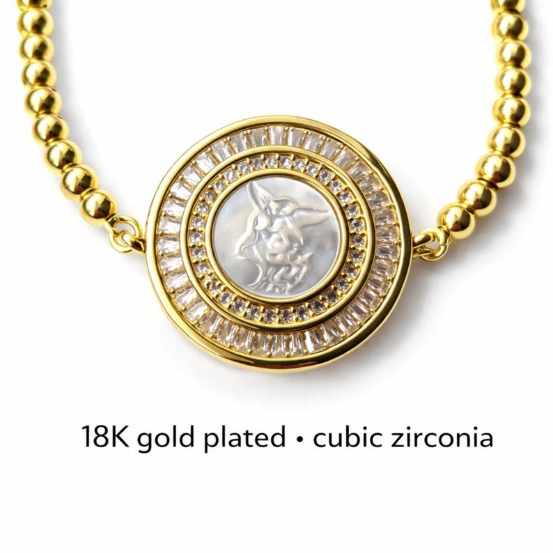 May include: Gold-plated bracelet with a round pendant featuring a carved design and cubic zirconia accents. The pendant has a central design and is surrounded by a ring of clear stones. The bracelet is made of small, gold-colored beads. Text on the image reads: "18K gold plated &bull; cubic zirconia."