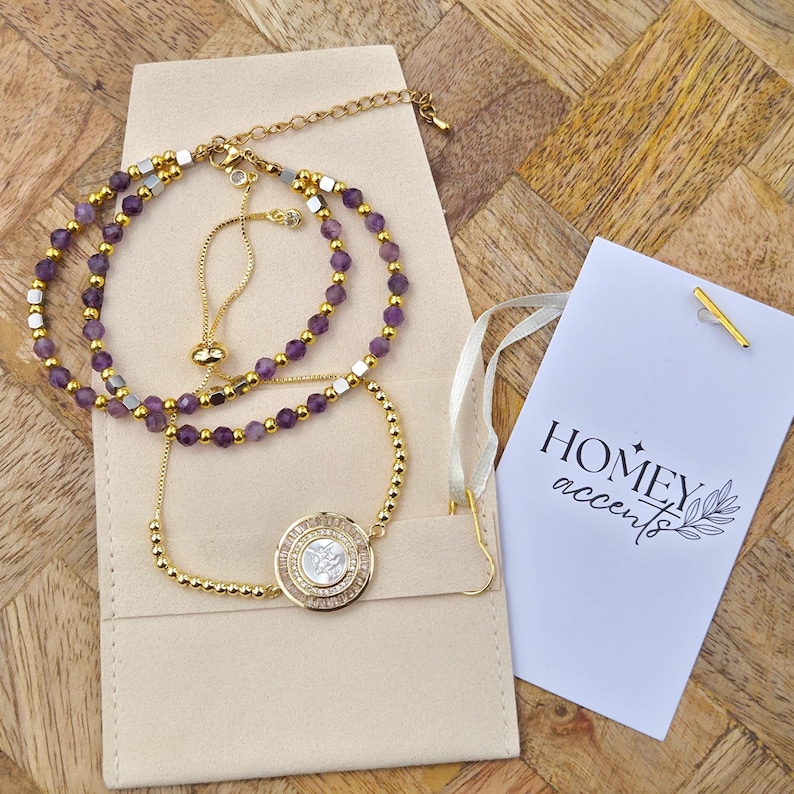 May include: Gold-toned bracelets with purple beads and a round pendant with a white design are displayed on a beige pouch. A tag with the text "HOMEY accents" is also present. The bracelets are on a wooden surface.