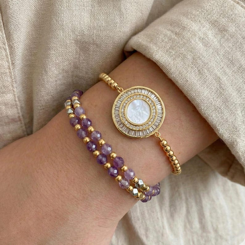 May include: Two bracelets on a wrist. One is gold with a circular pendant featuring a white center and surrounding clear stones. The other bracelet is made of purple beads and gold accents. The bracelets are on a beige background.
