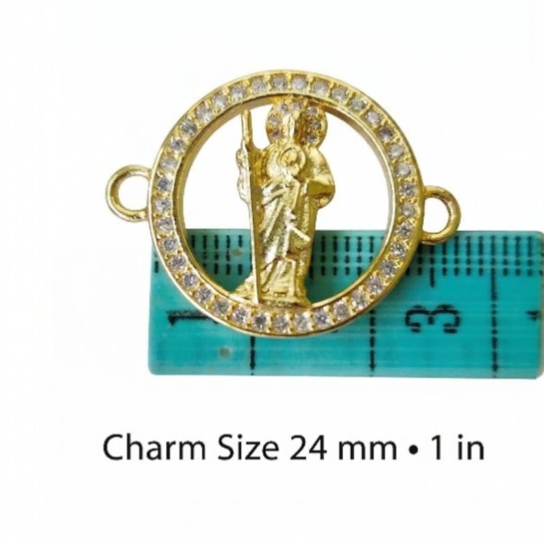May include: A gold-colored charm with a saint figure inside a circle, surrounded by small clear stones. The charm has two loops for attachment. The text "Charm Size 24 mm &bull; 1 in" is below the charm, which is placed on a teal ruler.