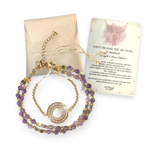 May include: A gold-toned bracelet with amethyst beads and a central circular charm. The bracelet is accompanied by a small beige pouch and a card with text about Saint Michael the Archangel and Amethyst.
