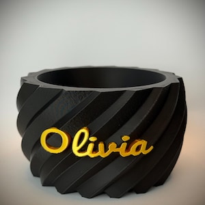 Op de afbeelding: Een zwarte, spiraalvormige plantenbak met de naam "Olivia" in gouden letters. De plantenbak heeft een uniek, gestructureerd ontwerp en een brede opening. Dit decoratieve item is geschikt voor kleine planten of als stijlvolle opbergcontainer.