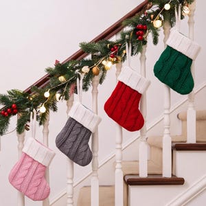May include: Four cable-knit Christmas stockings hanging from a staircase railing. The stockings are pink, gray, red, and green, each featuring a white ribbed cuff and a hanging loop. The knit pattern consists of vertical columns of twisted cables.