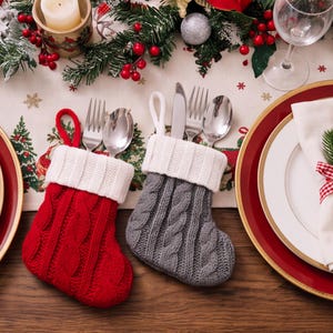 May include: Two miniature cable-knit stockings, one red and one gray, each featuring a white ribbed cuff and a hanging loop. The stockings serve as holders for stainless steel silverware, including forks, spoons, and knives, placed on a table.