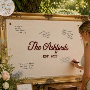 May include: A large rectangular wedding guest book sign in a gold ornate frame with a cream fabric center. The text reads The Ashfords EST. 2027 in dark red script and block letters, surrounded by handwritten congratulatory notes in black ink.