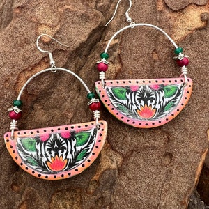 May include: A pair of handcrafted earrings with a half-circle design. The earrings feature a pink and orange painted surface with a floral and zebra stripe pattern. Silver-tone metal hoops and beaded accents in red and green complete the design.
