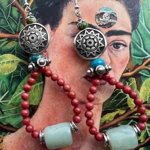 May include: A pair of long, silver earrings with turquoise and red beads. The earrings feature a large, silver charm with a starburst design. The earrings are hanging against a background of a painting of a woman.