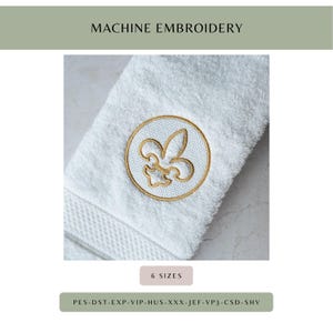 May include: White terry cloth towel with a gold embroidered fleur-de-lis emblem. The emblem is circular and features a detailed design. The words "MACHINE EMBROIDERY" are at the top. The towel has a textured border.