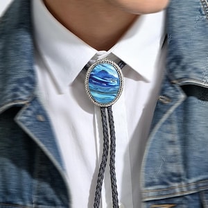 May include: A bolo tie featuring an oval-shaped centerpiece with a swirling blue and white marbled pattern, encased in a silver-toned braided metal frame. The cord is made of dark grey braided material, worn over a white collared shirt and denim jacket.