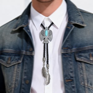 May include: Silver bolo tie featuring an eagle pendant with a turquoise stone center. The pendant hangs from a black braided cord, accented with silver feather charms. The bolo tie is worn over a white collared shirt and a denim jacket.